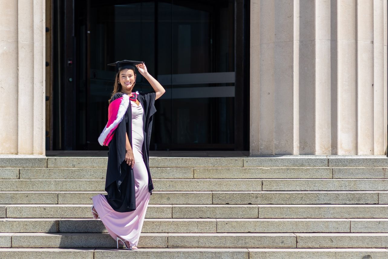 Graduate in black gown and pink hood poses proudly on university steps in academic cap and gown.