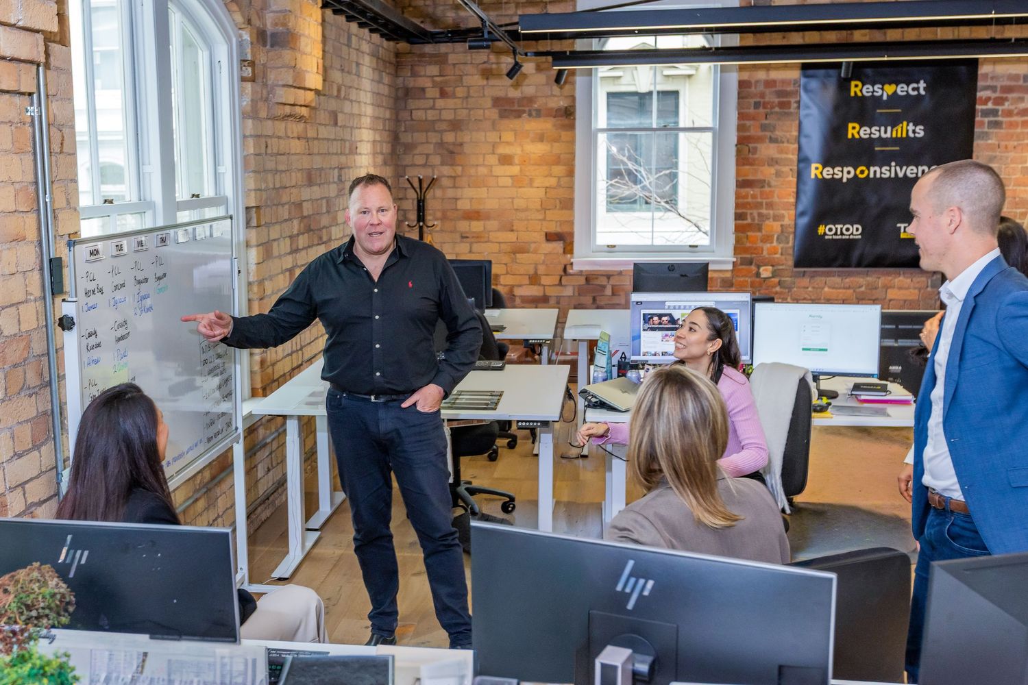 Professional team meeting in modern brick-walled office with presenter at whiteboard and colleagues at HP computers.