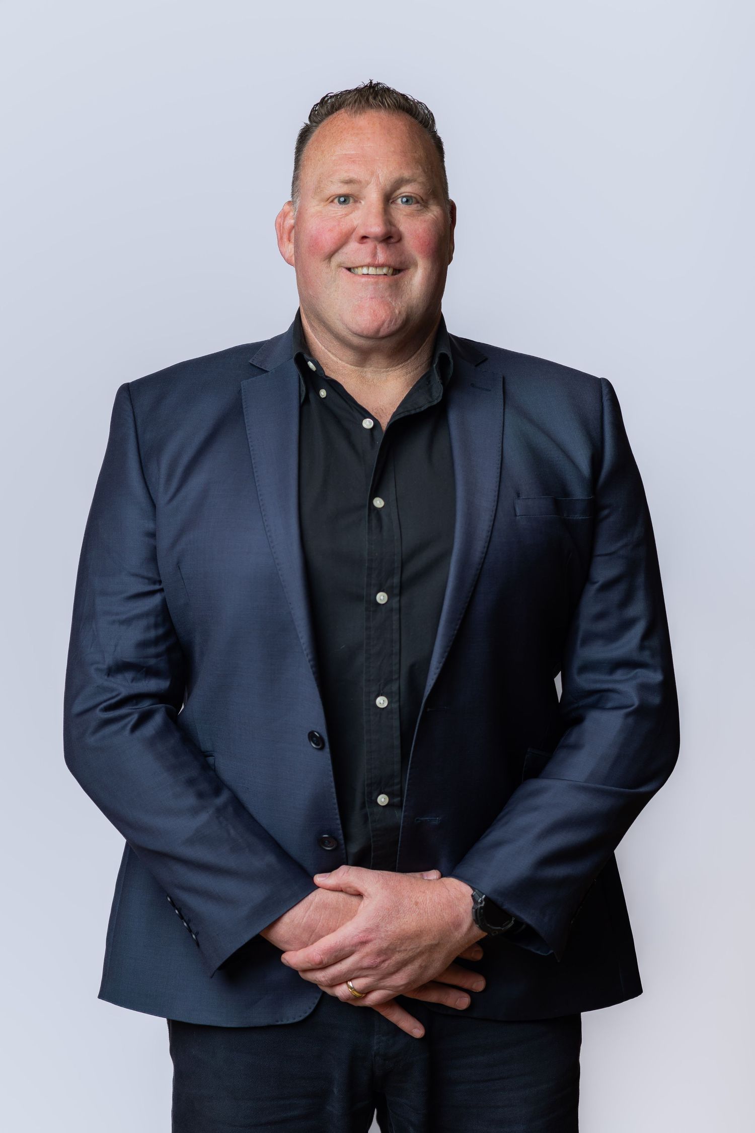 Smiling man in navy blue blazer and black shirt posing with hands clasped against light gray background.