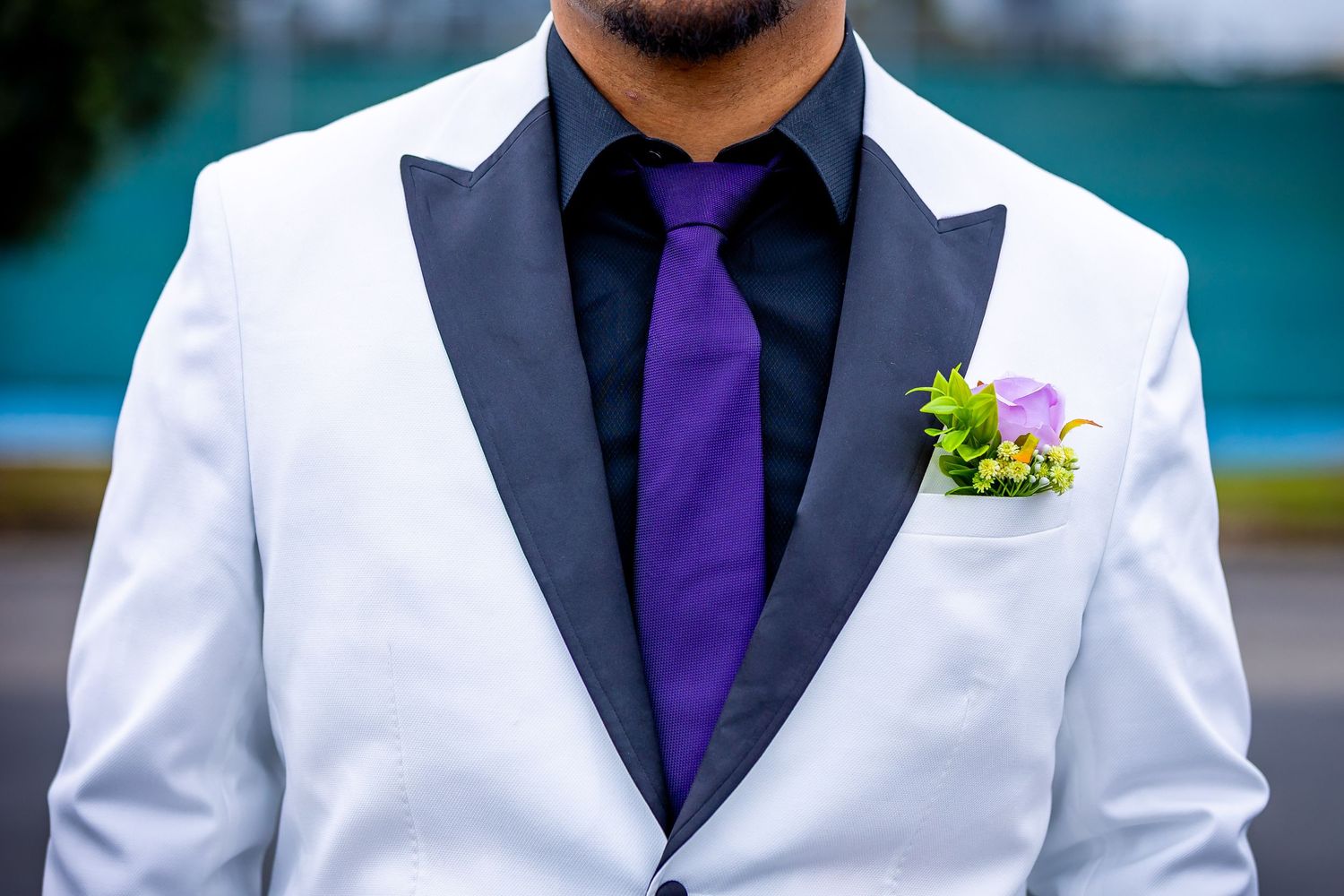Man in white tuxedo jacket with black lapels, purple tie, and floral boutonniere at formal event.
