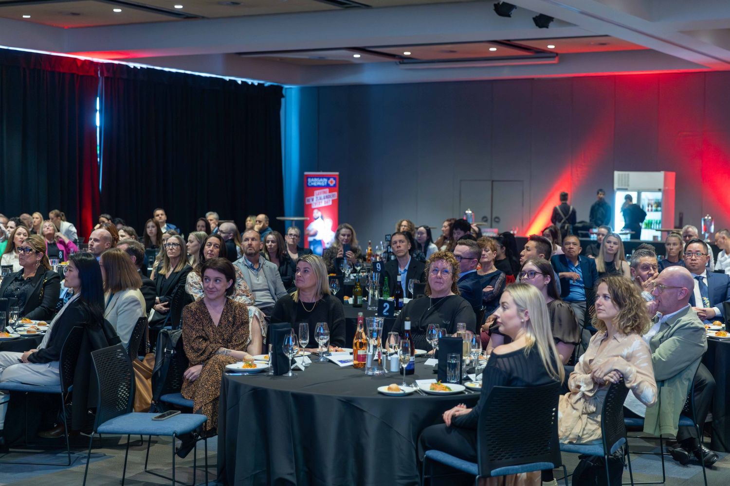 Crowded business networking event with attendees seated at round tables under mood lighting.