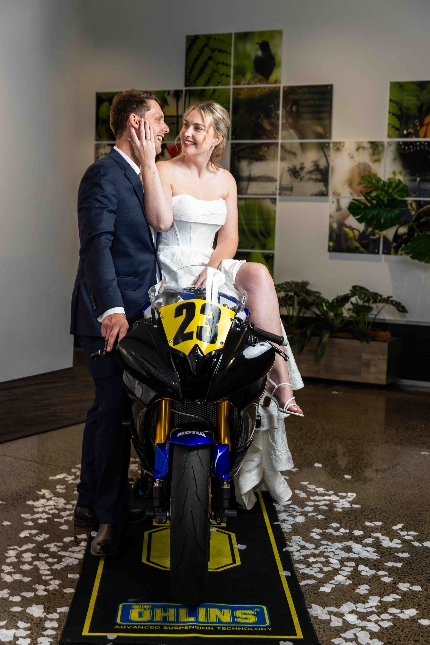 Wedding couple posing with racing motorcycle surrounded by flower petals indoors.
