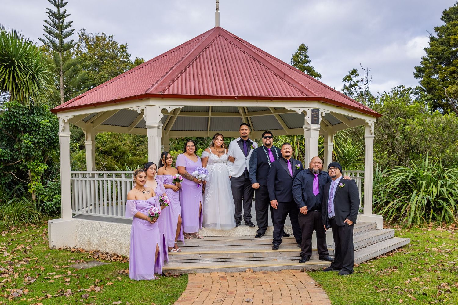 Wedding party in purple and white attire poses before a white gazebo with red roof in a lush garden setting.