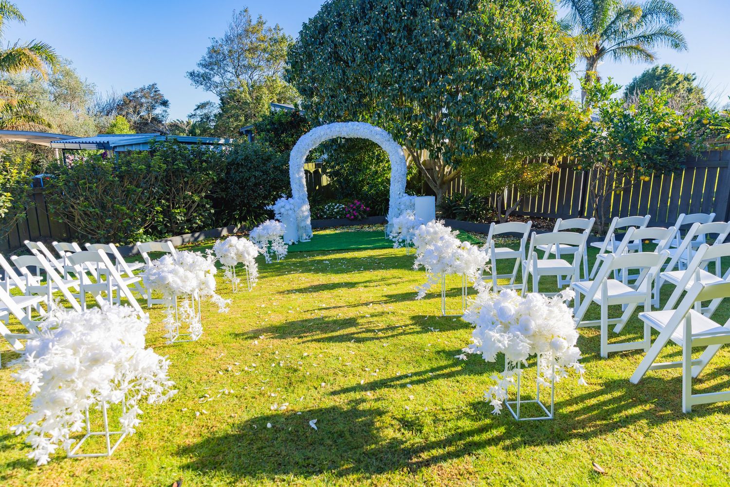 Outdoor wedding ceremony setup with white chairs, floral arrangements, and a white arch on a green lawn.