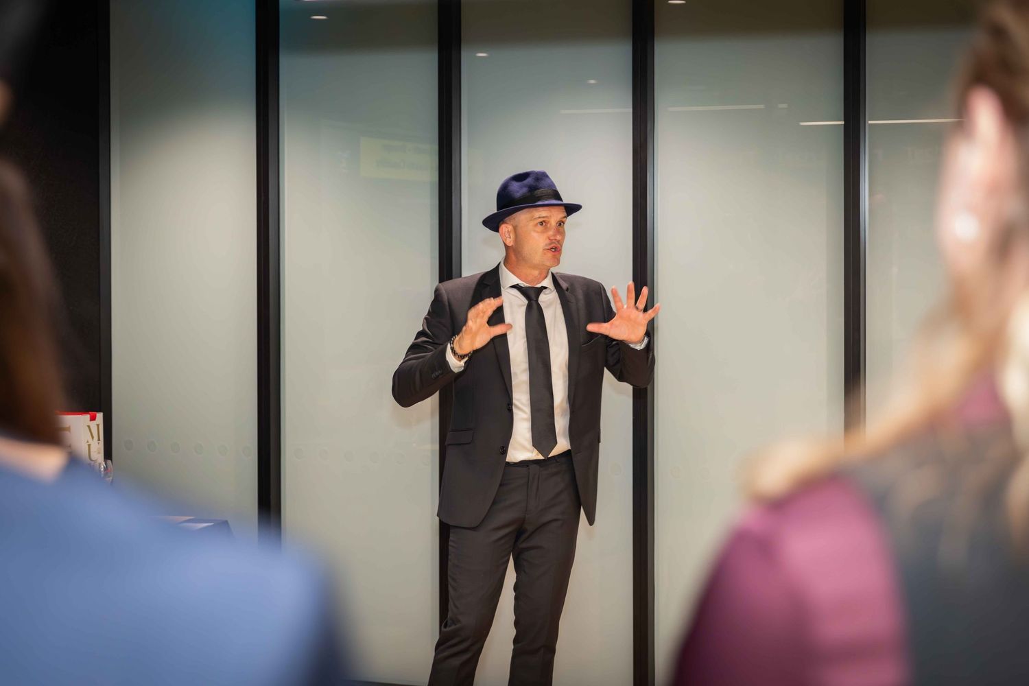 Man in black suit and blue hat gesturing while speaking to an audience in a modern office setting.