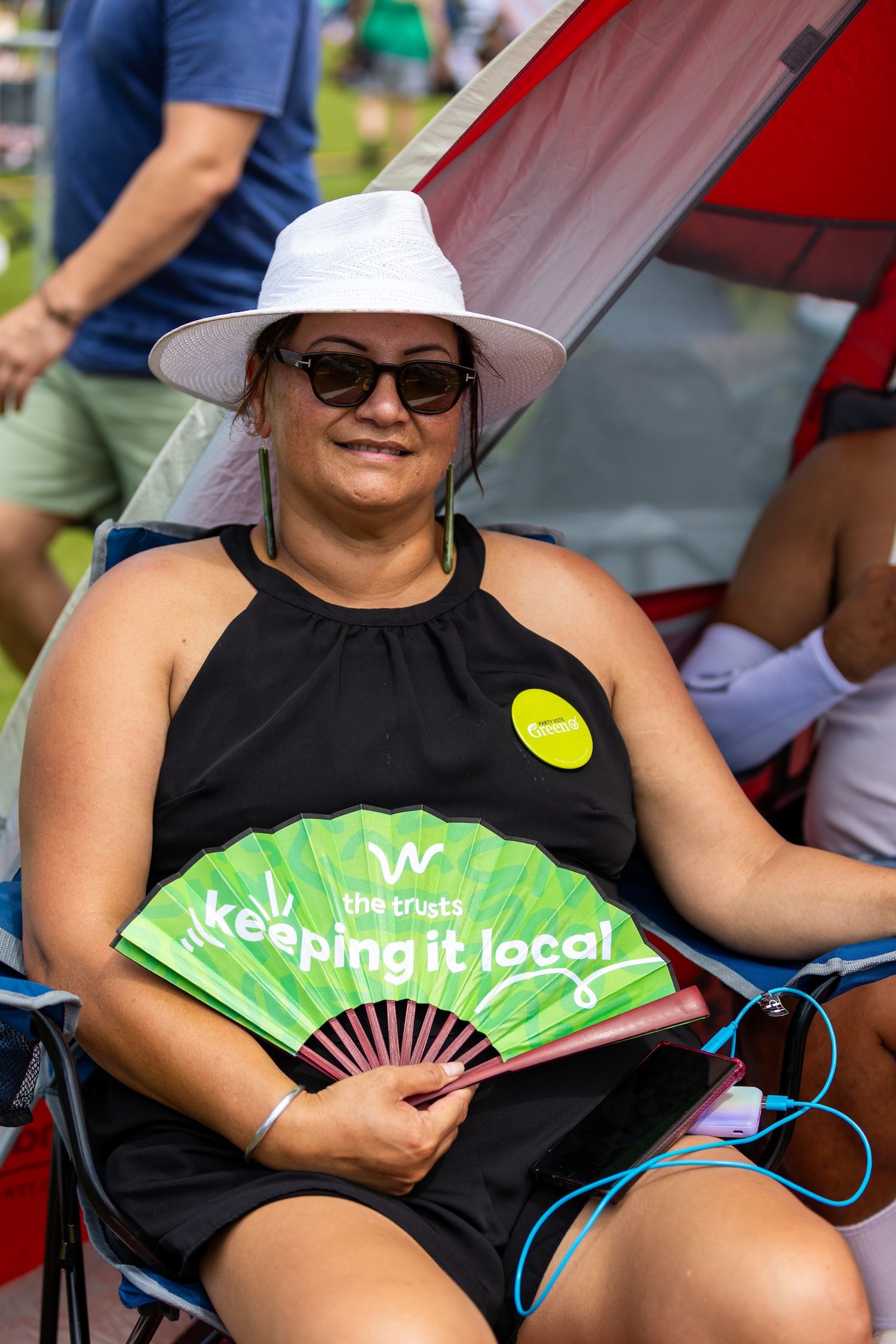 People relax in red camping chairs while using green folding fans to stay cool.