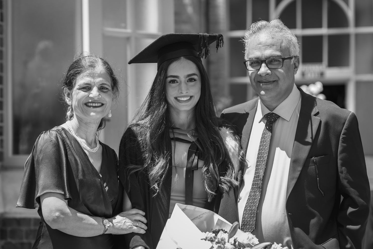 Black and white photo of a graduate in cap and gown posing with two proud family members at graduation ceremony.