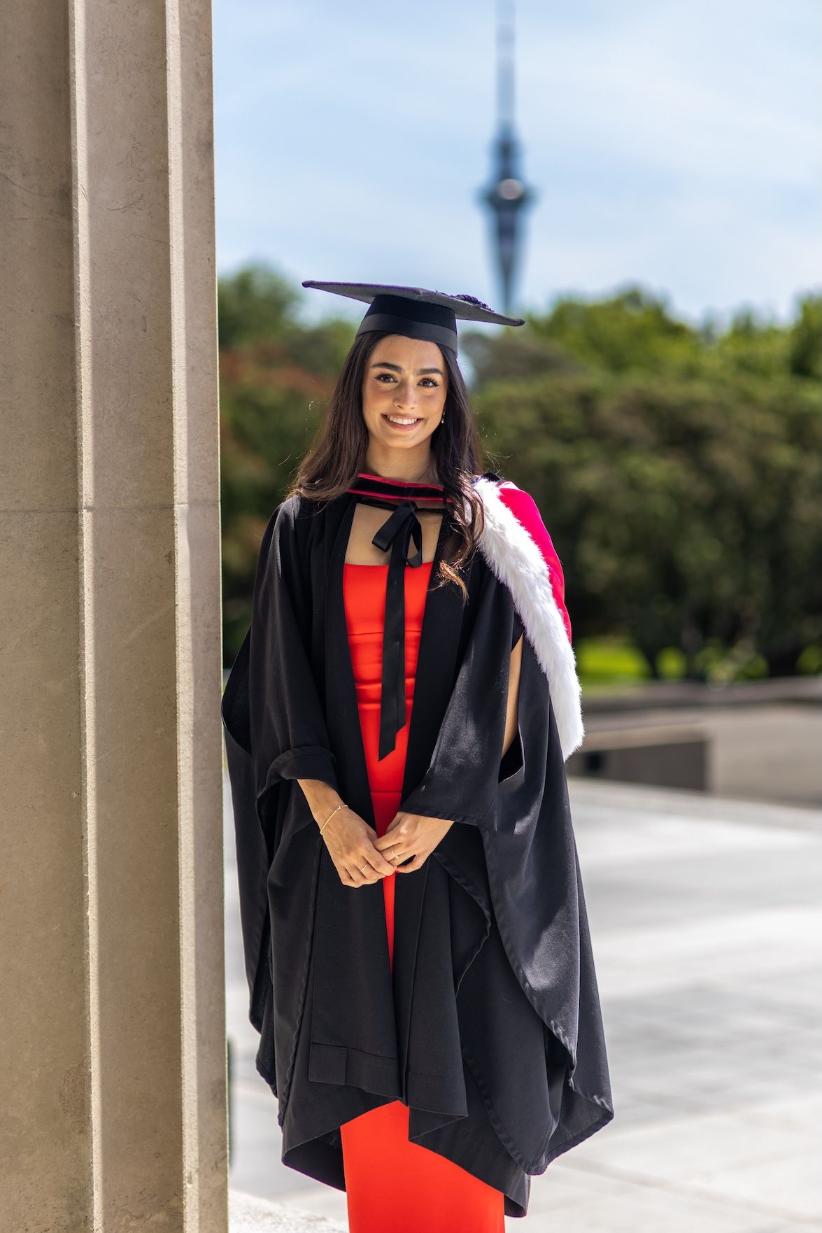 Smiling female graduate in black gown and mortarboard cap poses by a column with a tower in the background.