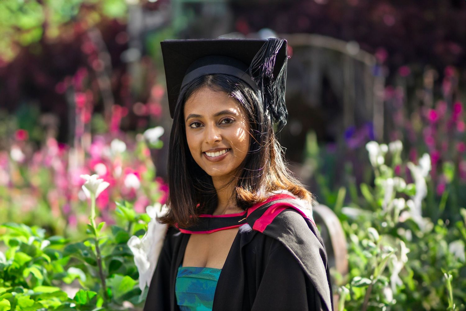Smiling female graduate in cap and gown posing outdoors among colorful flowers on graduation day.