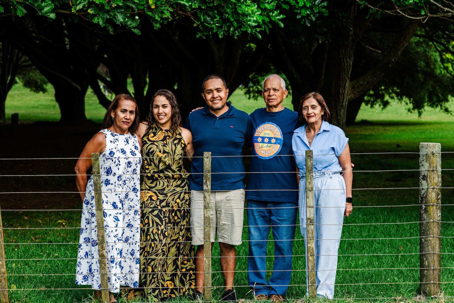 Five people smiling together outdoors near a wire fence surrounded by lush green trees.