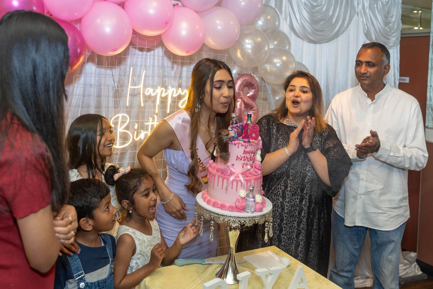 Family celebrating a birthday with a pink decorated cake, balloons, and Happy Birthday sign at a festive indoor party.