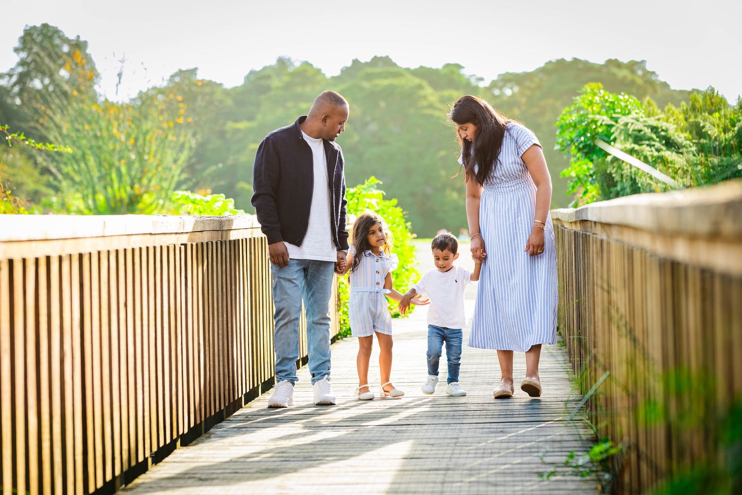Family of four walking on a wooden bridge surrounded by lush greenery on a sunny day.