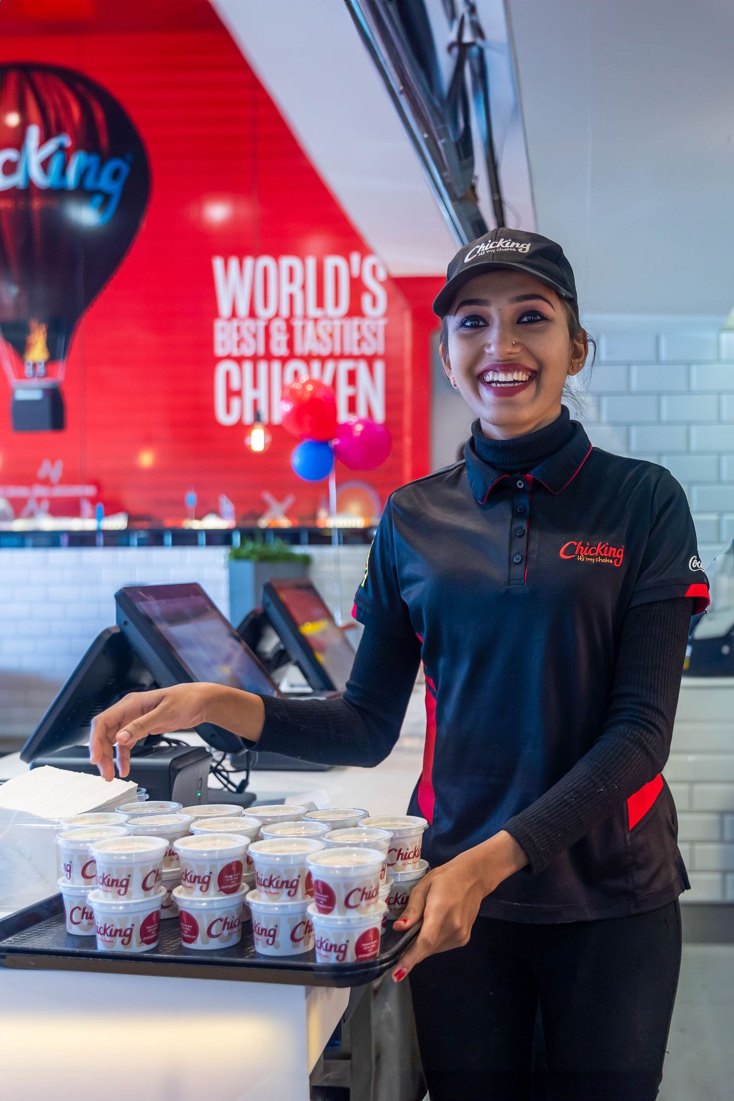 Smiling Chicking employee holding a tray of cups at a fast food counter with World's Best & Tastiest Chicken sign.