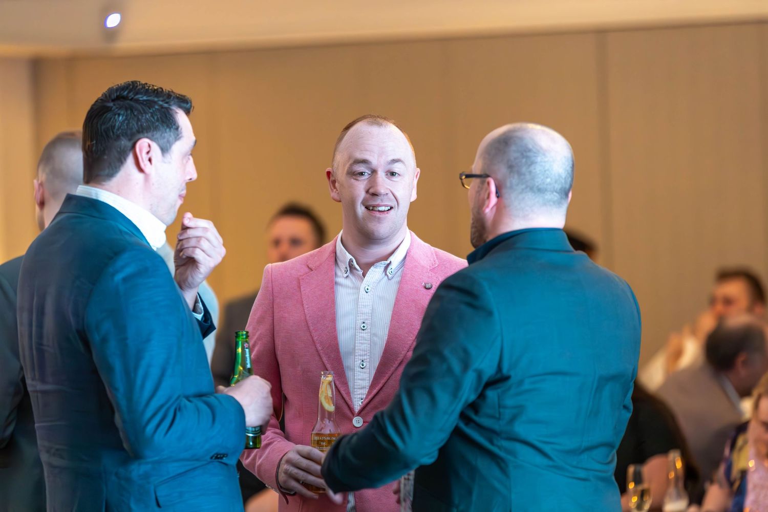 Three men in suits socializing at a formal event, with the center man smiling in a pink blazer.