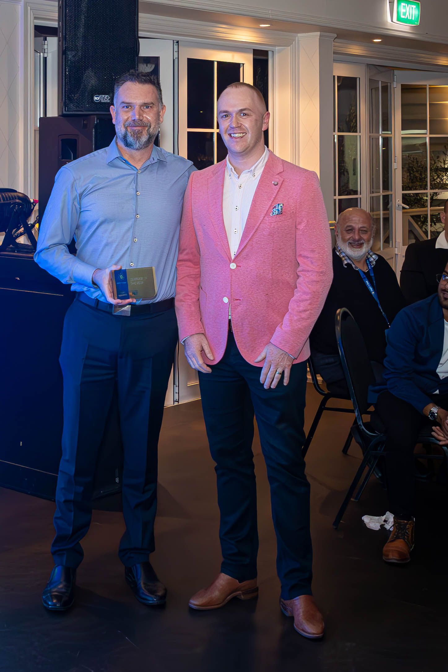 Two men pose together at an awards ceremony, one holding a plaque, dressed in smart casual attire.