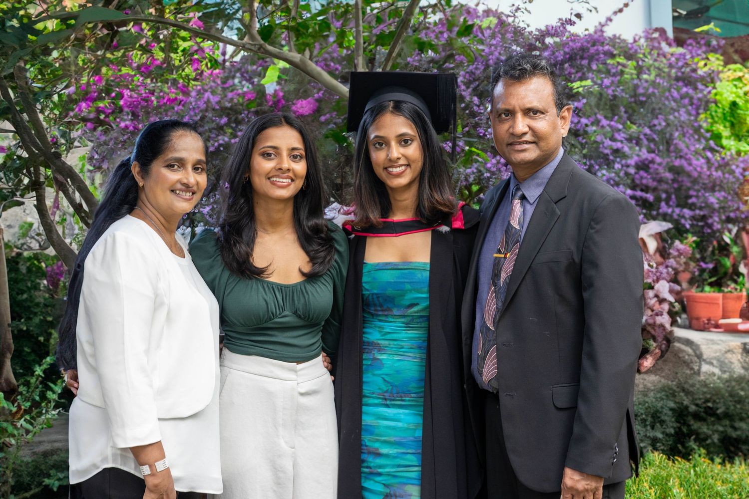 Family of four posing together at a graduation ceremony outdoors with purple flowering trees in the background.