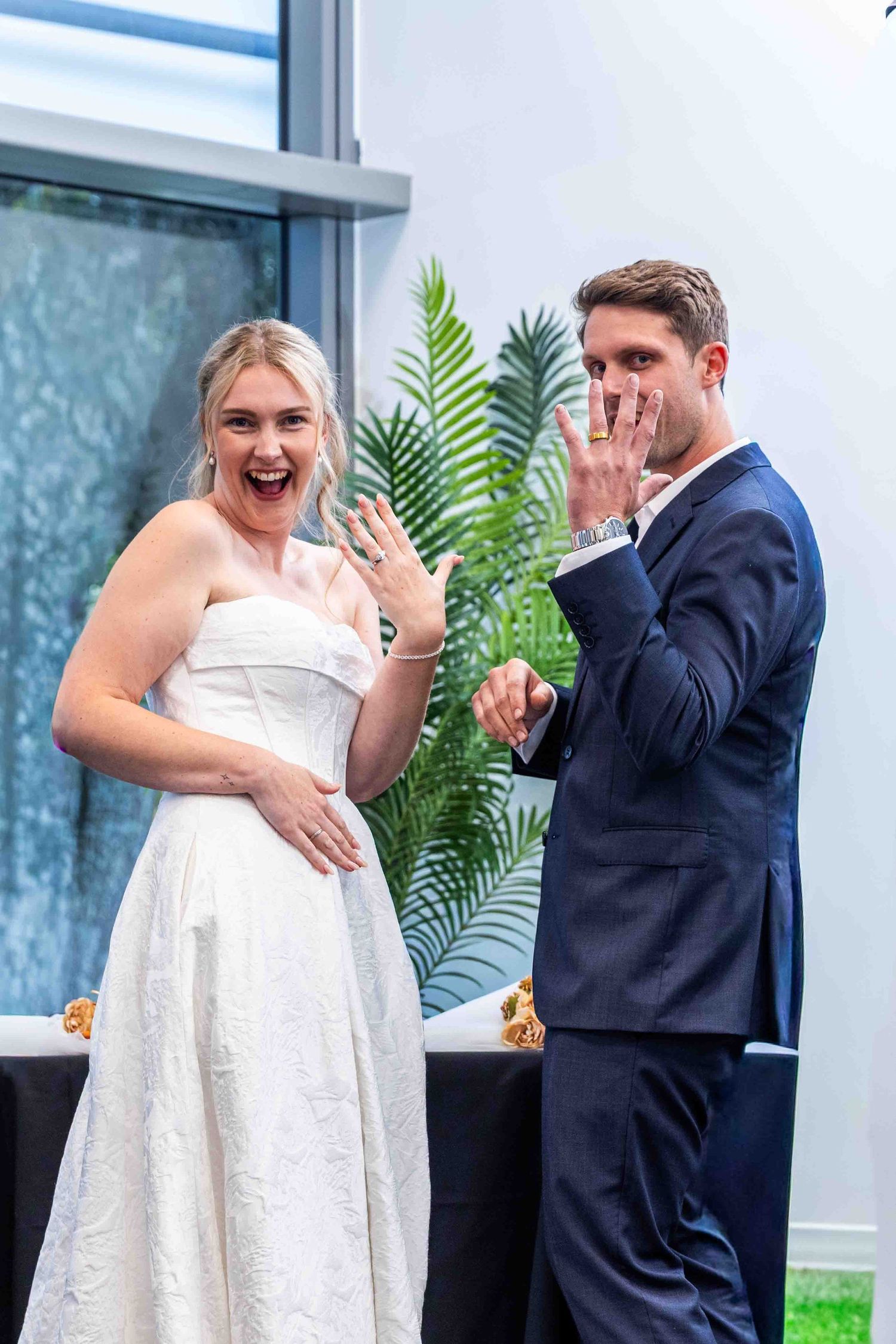 Couple shares playful moment during wedding photoshoot with tropical plant decor and modern building backdrop.