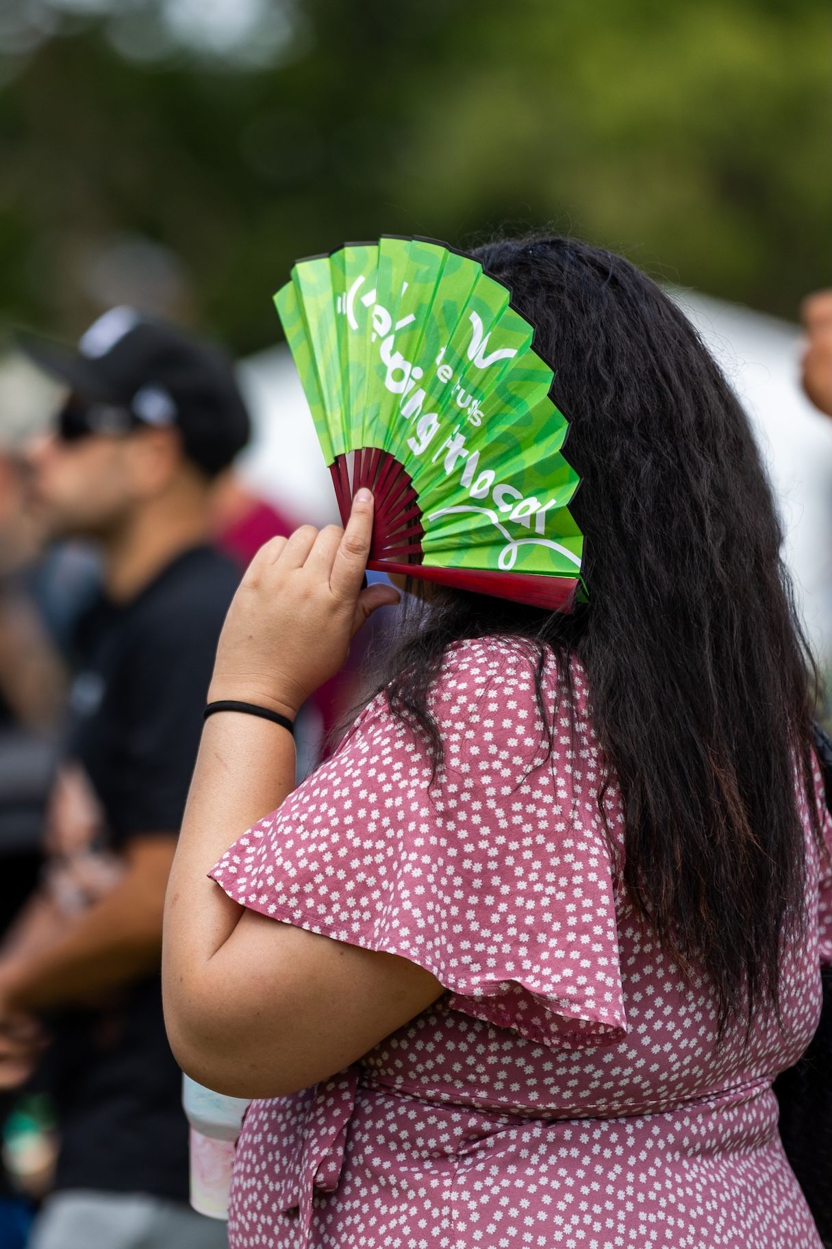 Person in pink floral dress holding bright green decorative fan at outdoor event.