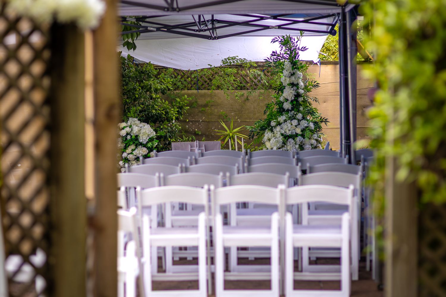Elegant outdoor wedding ceremony setup with white chairs under a canopy, adorned with floral arrangements and lush greenery.