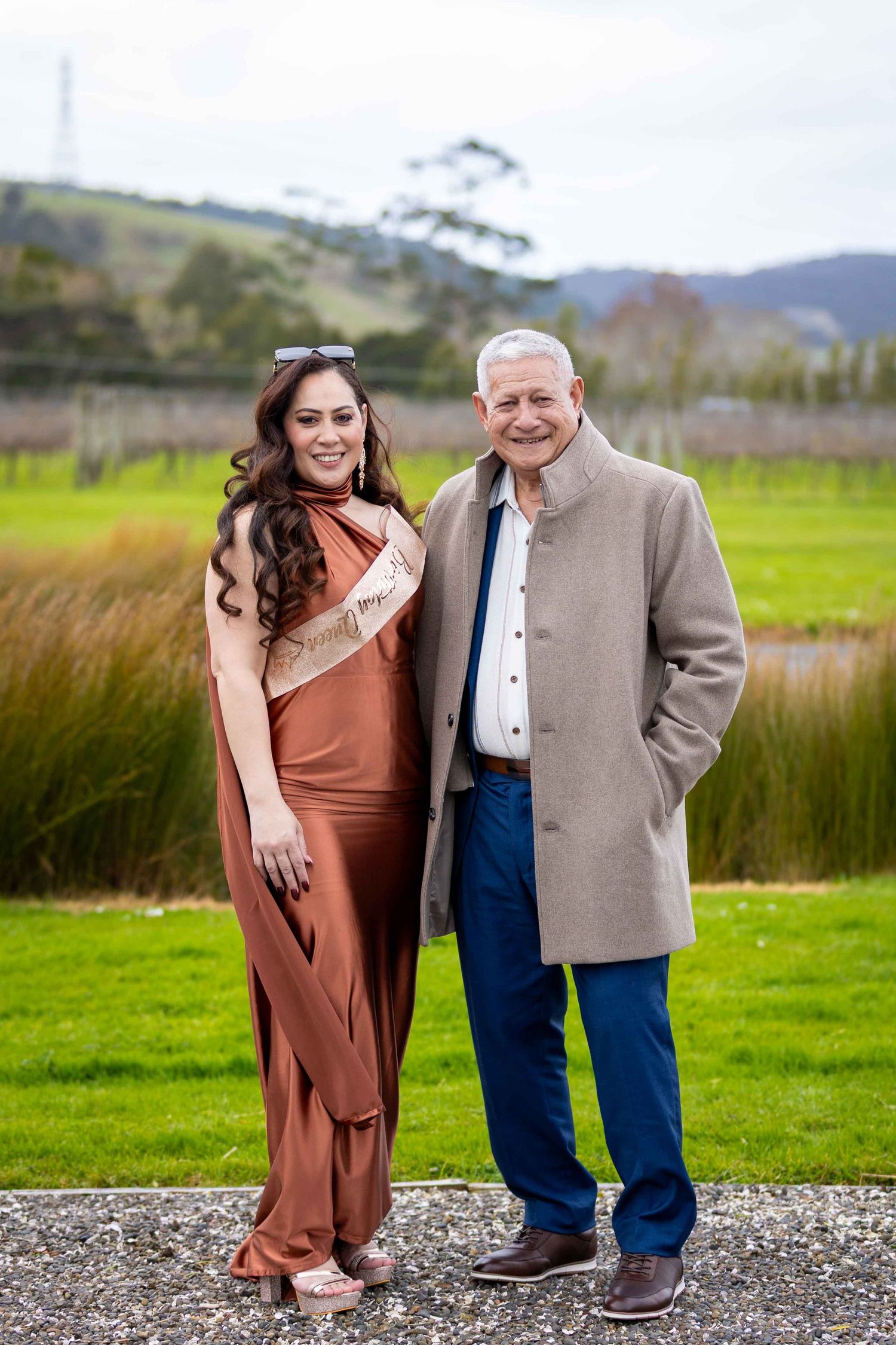 A woman in a rust sari and a man in a beige coat pose together outdoors at a vineyard.