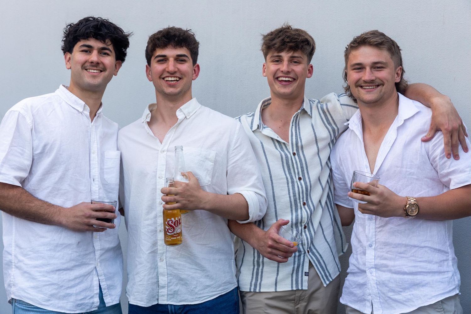 Four young men in white shirts smiling and holding drinks outdoors at a casual social gathering.