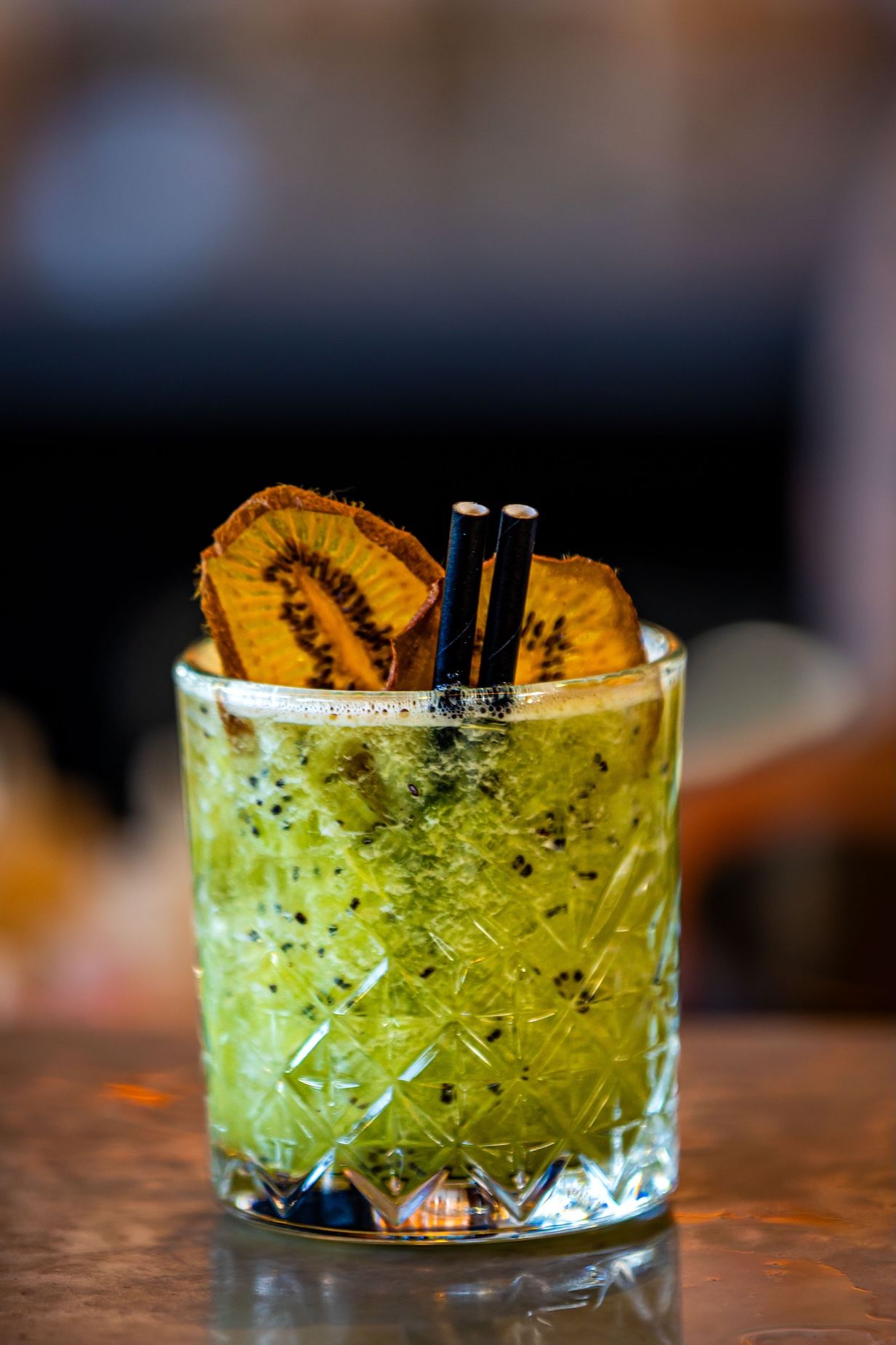 A refreshing green cocktail garnished with dried pineapple slices and black straws in a crystal glass on a wooden bar.