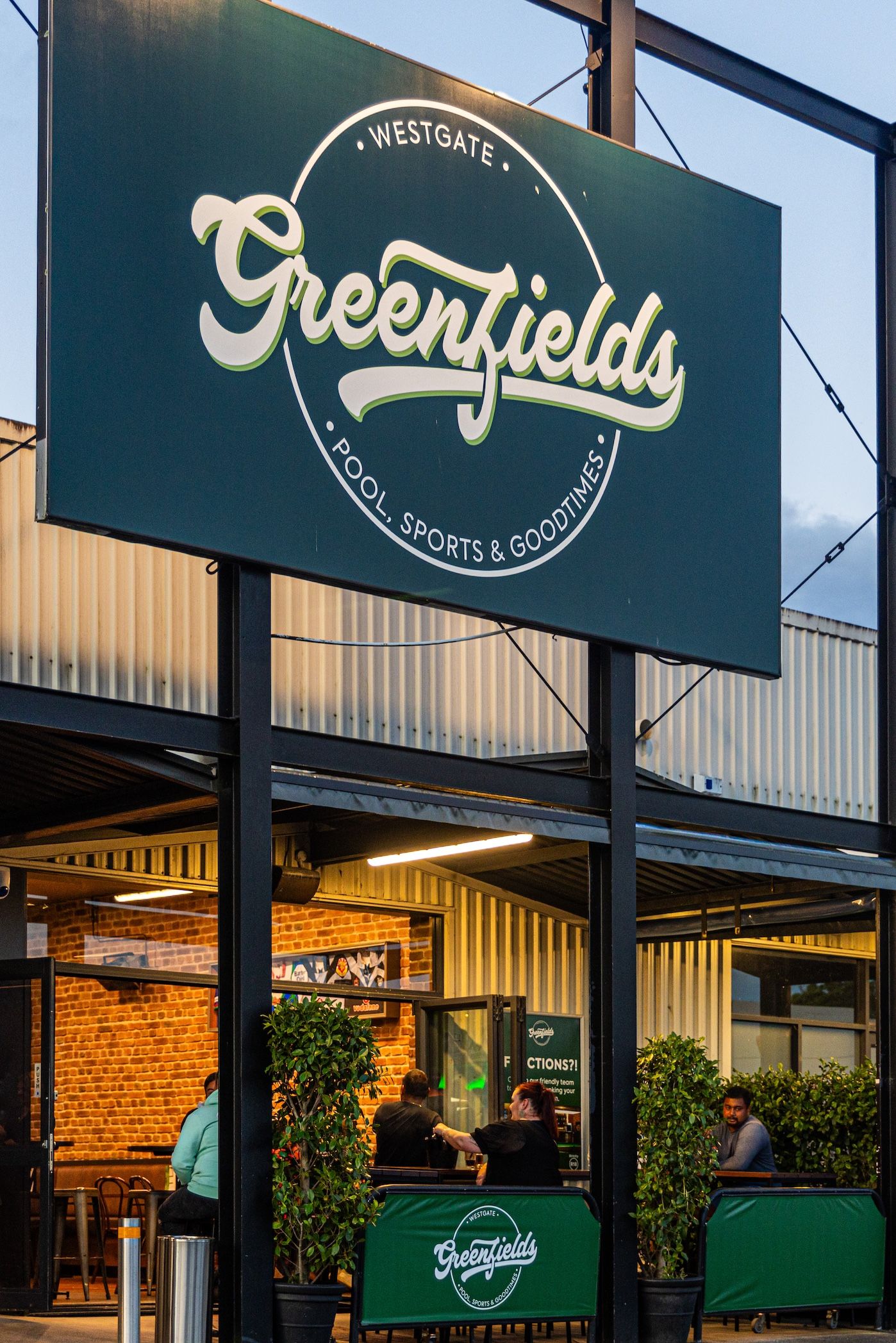Green storefront signage for Westgate Sports and Social with brick facade and decorative plants at dusk.