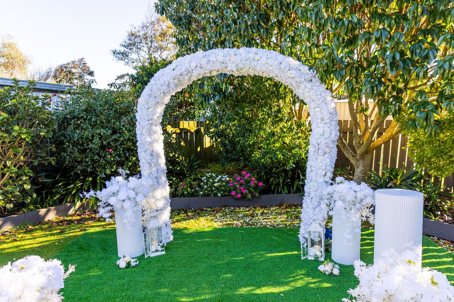 White floral arch wedding ceremony backdrop on lush green lawn surrounded by trees and white cylinder stands.