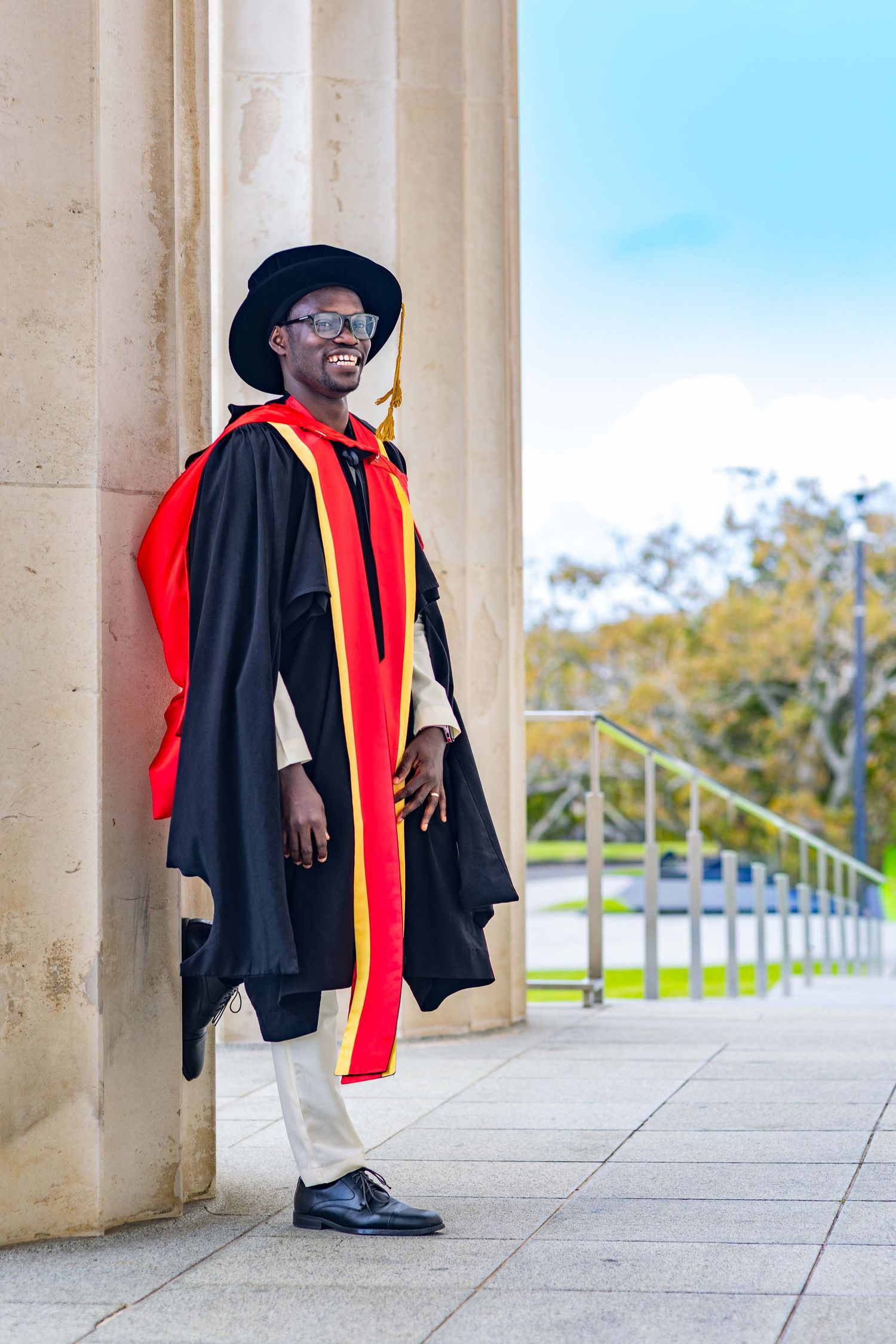 Smiling graduate in black and red doctoral gown and mortarboard hat posing between columns on graduation day.