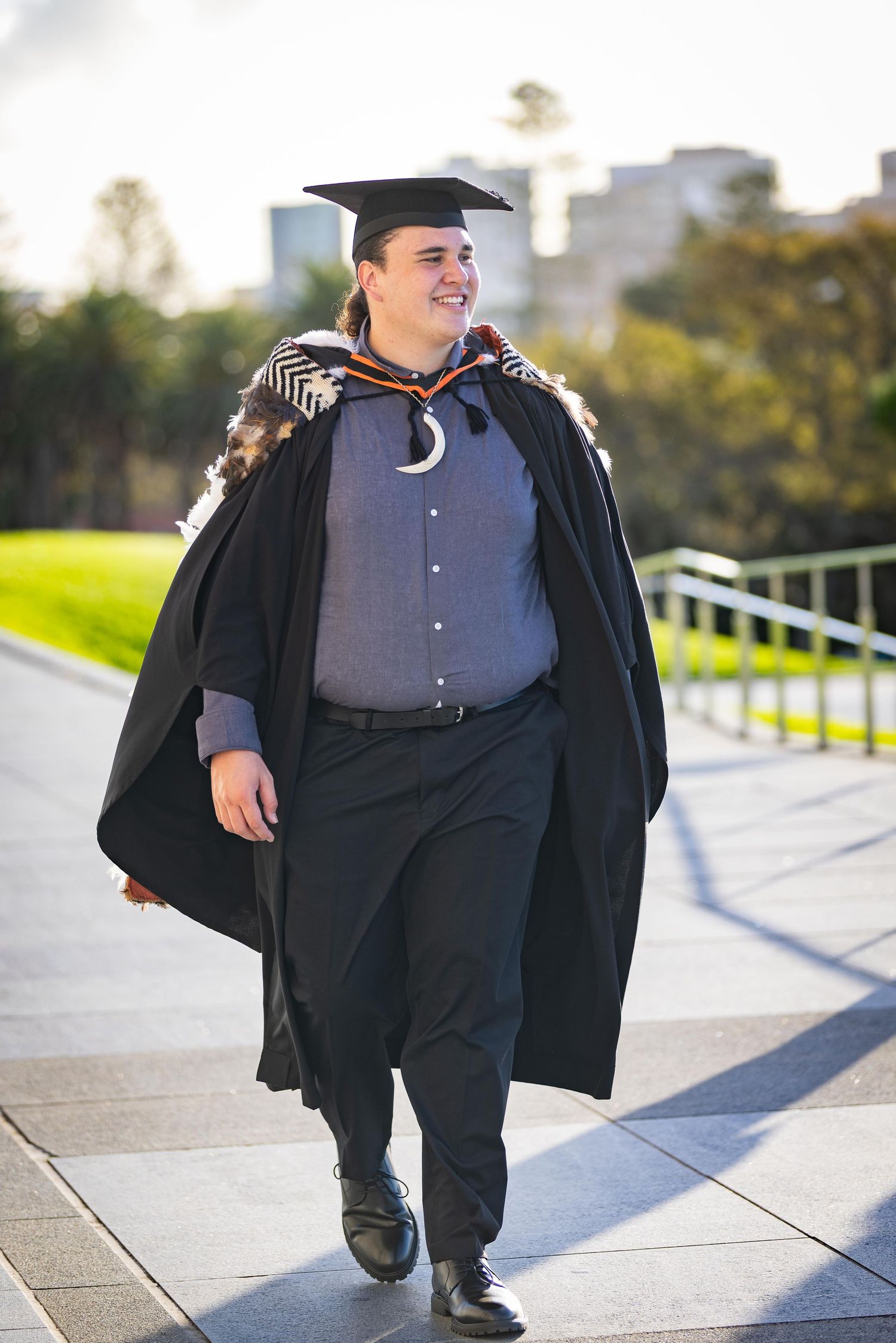 Graduate in black cap and gown smiles while walking outdoors on a sunny day at a university campus.