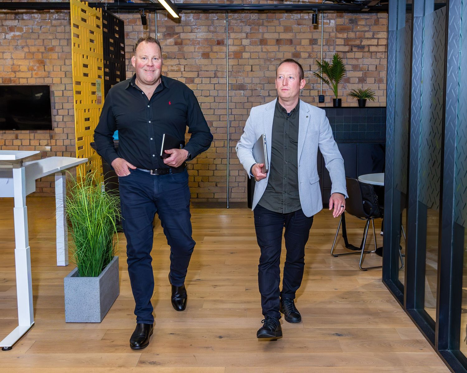 Two businessmen walking confidently through a modern brick-walled office with wooden floors and green plants.
