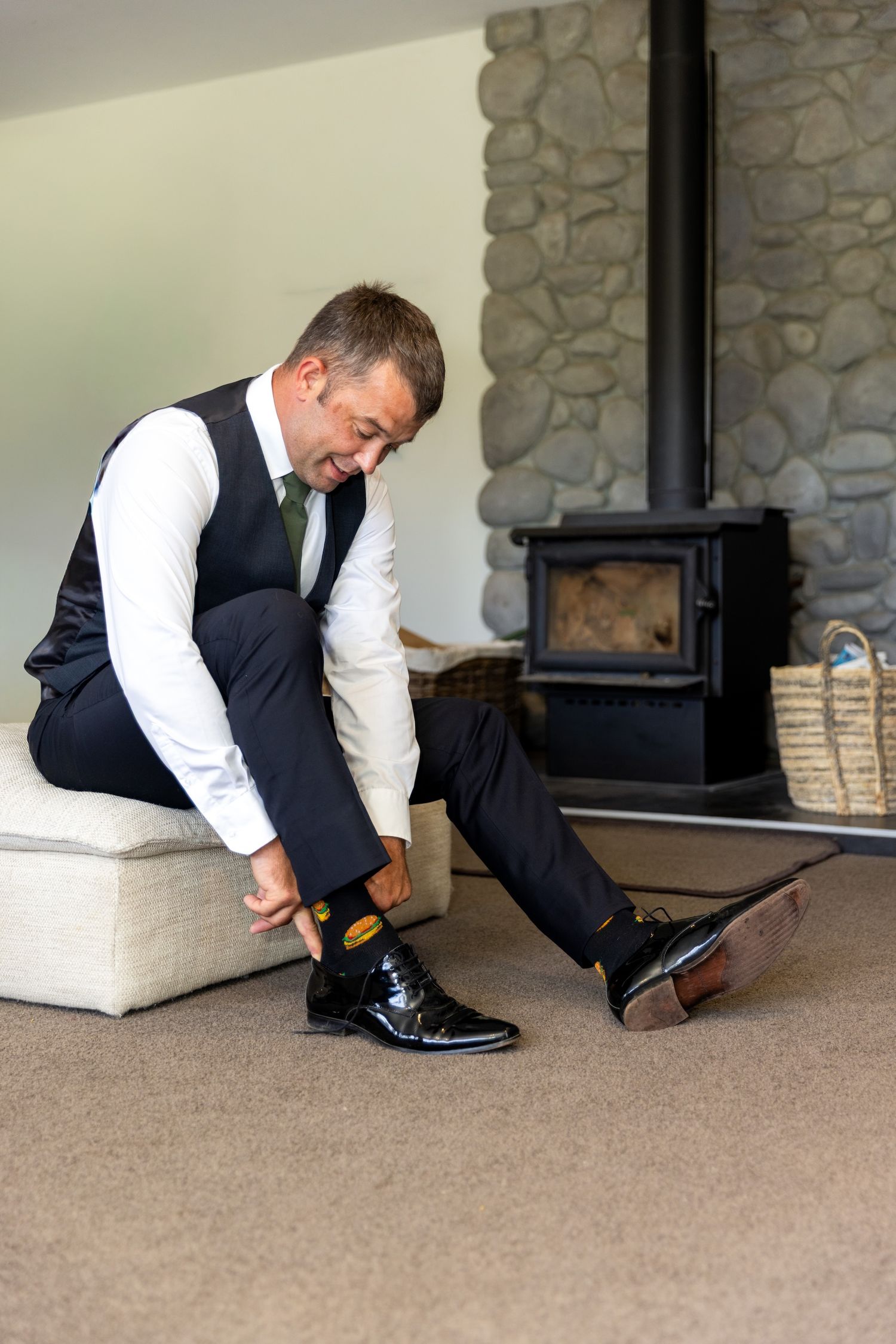 Man in formal vest and dress pants putting on black patent leather shoes near a stone fireplace.