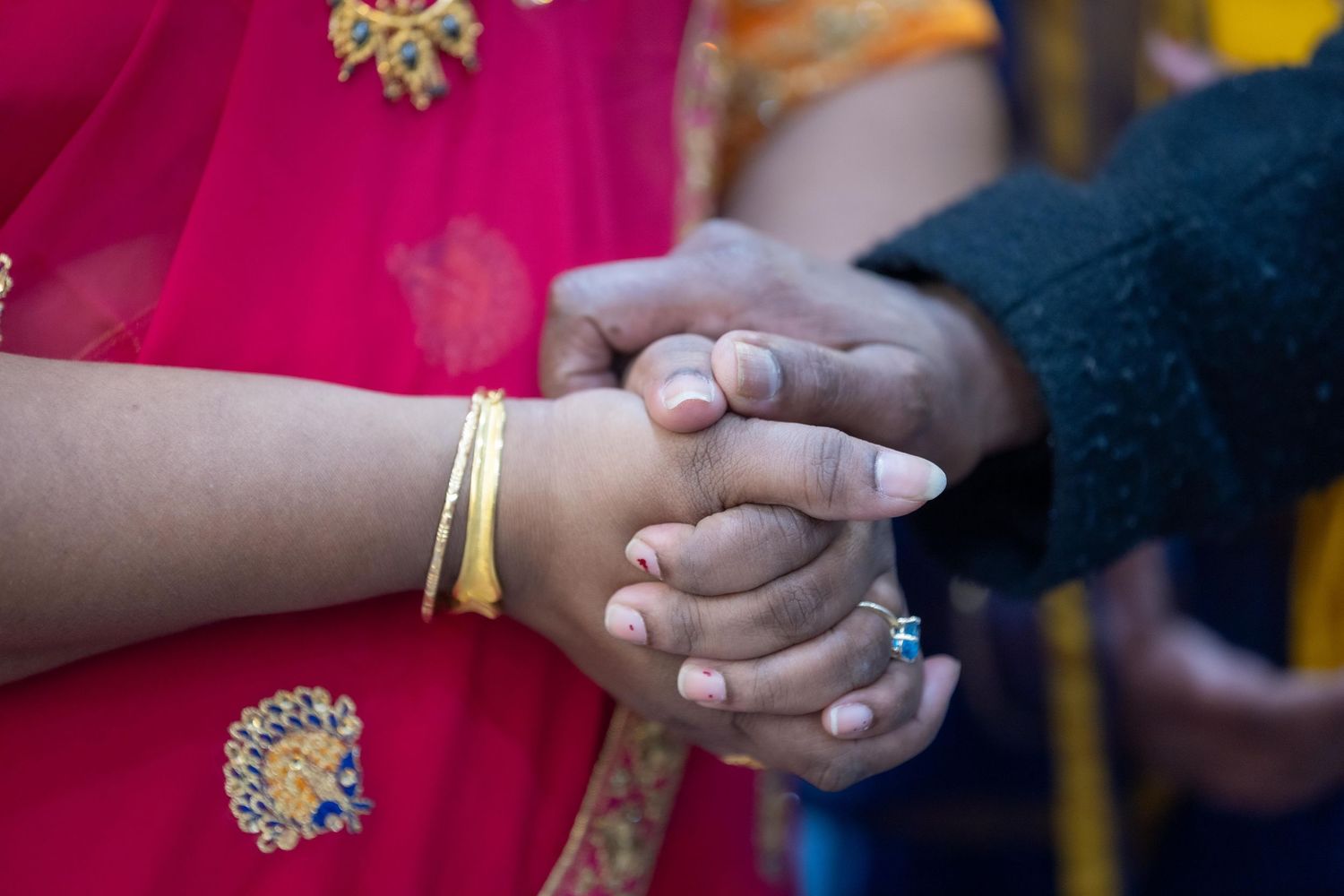 Two hands clasped together, one wearing gold bangles and a pink sari, in a traditional South Asian cultural ceremony.