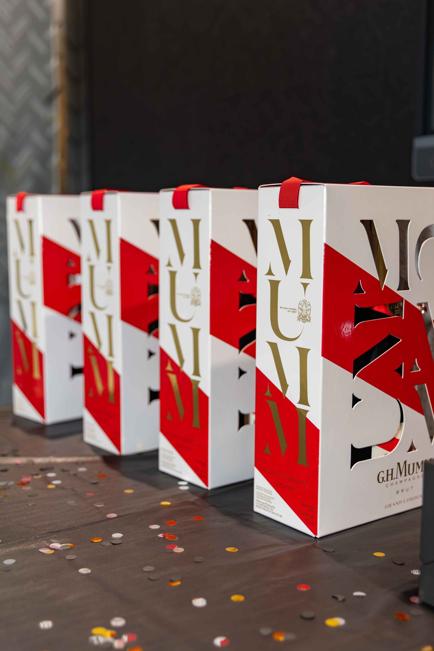 Row of G.H. Mumm champagne gift boxes with red and white design displayed on a table with confetti.