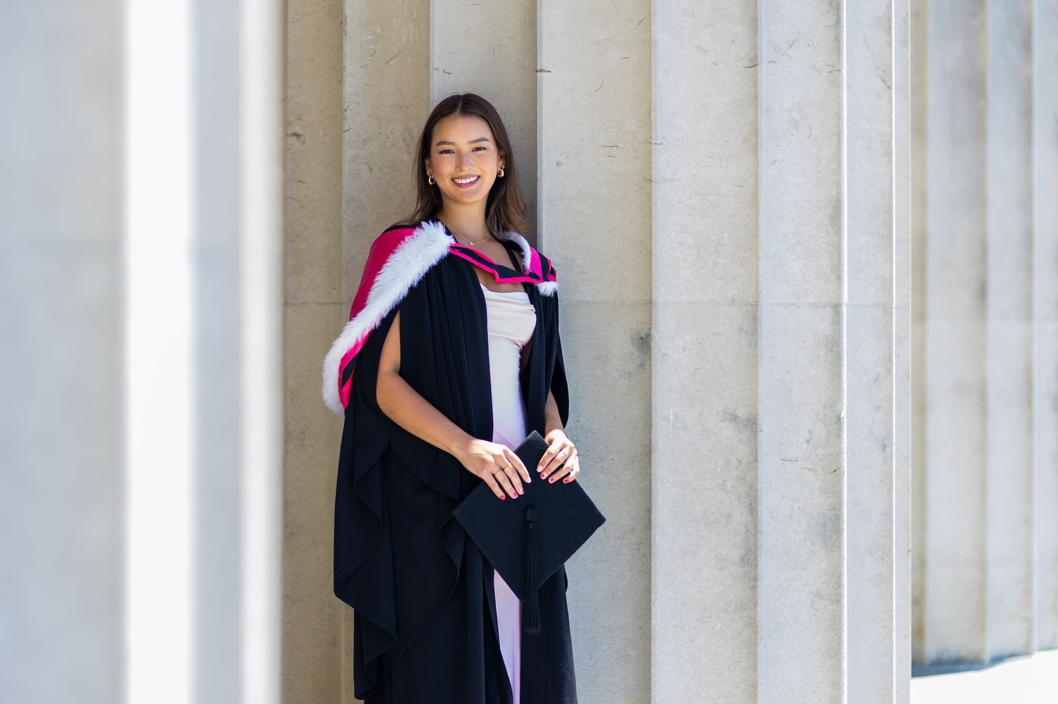 Young woman in black graduation gown with pink hood holds cap, smiling between stone columns.