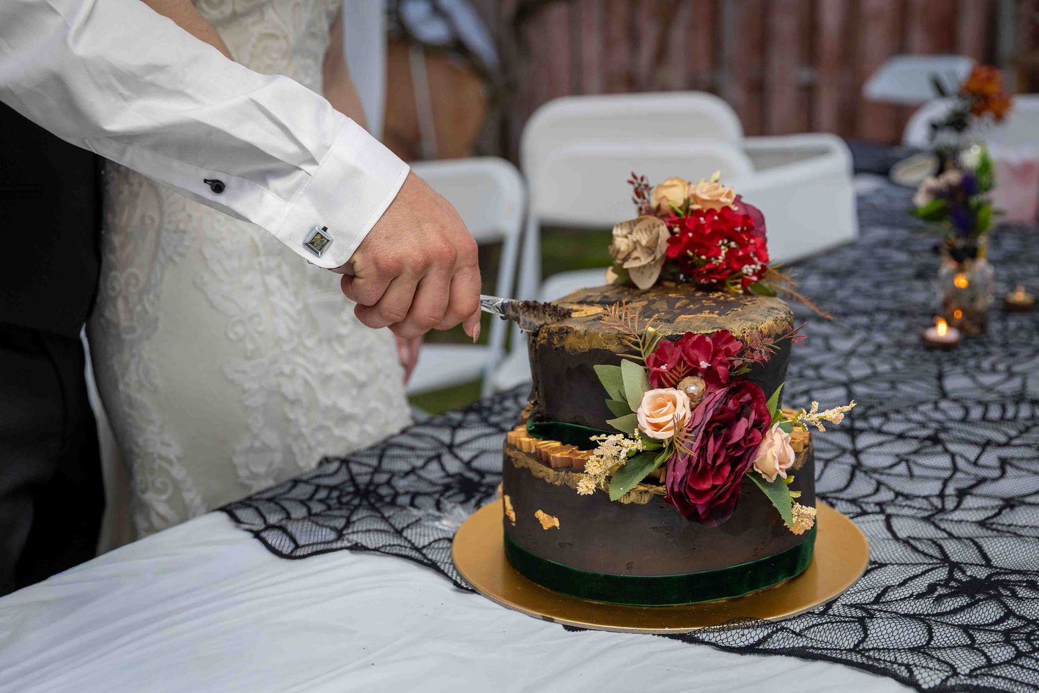 Bride and groom cutting a two-tier chocolate wedding cake decorated with red and pink flowers on a black lace tablecloth.