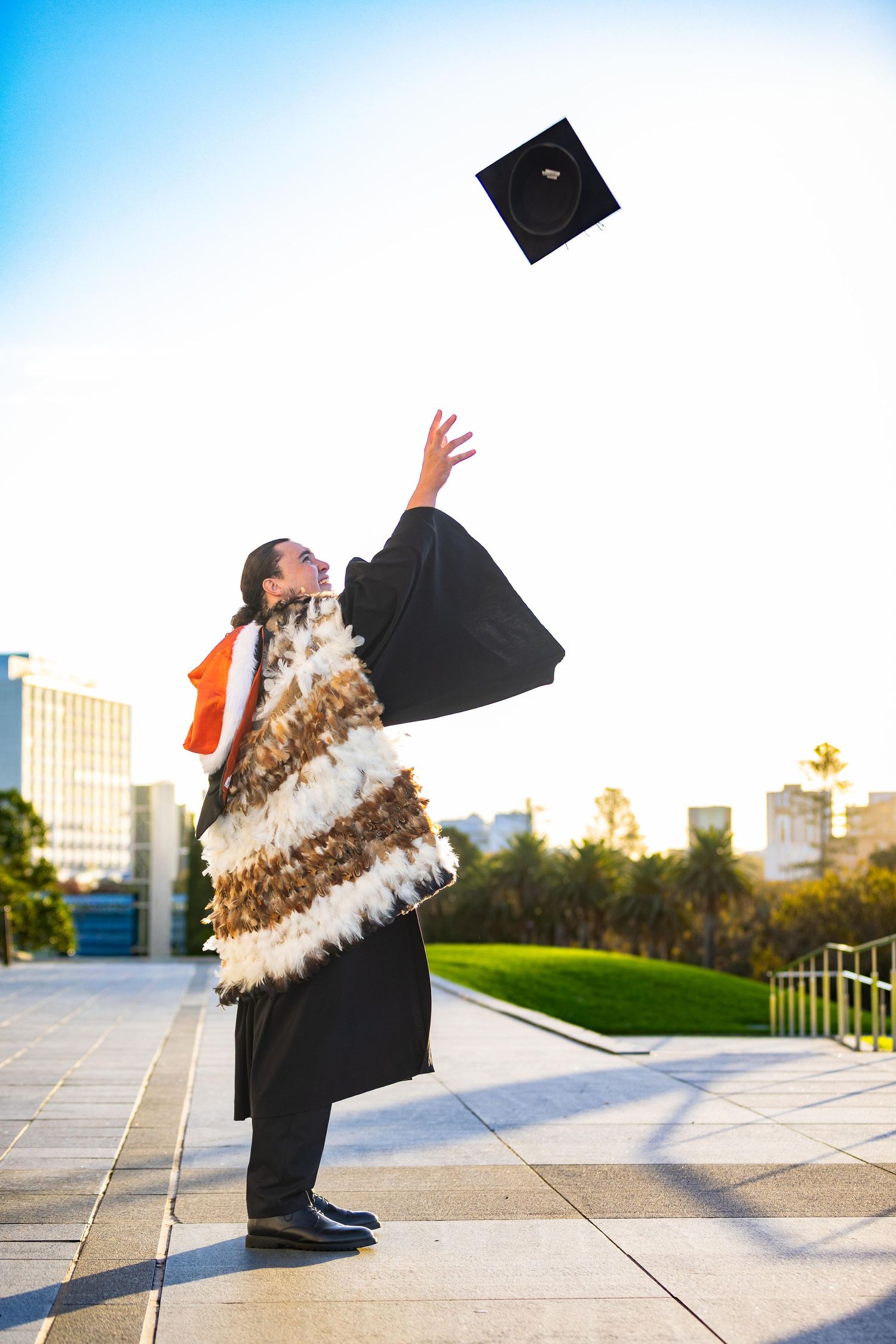 Graduate in black gown and traditional Maori cloak tosses mortarboard cap into the air outdoors.