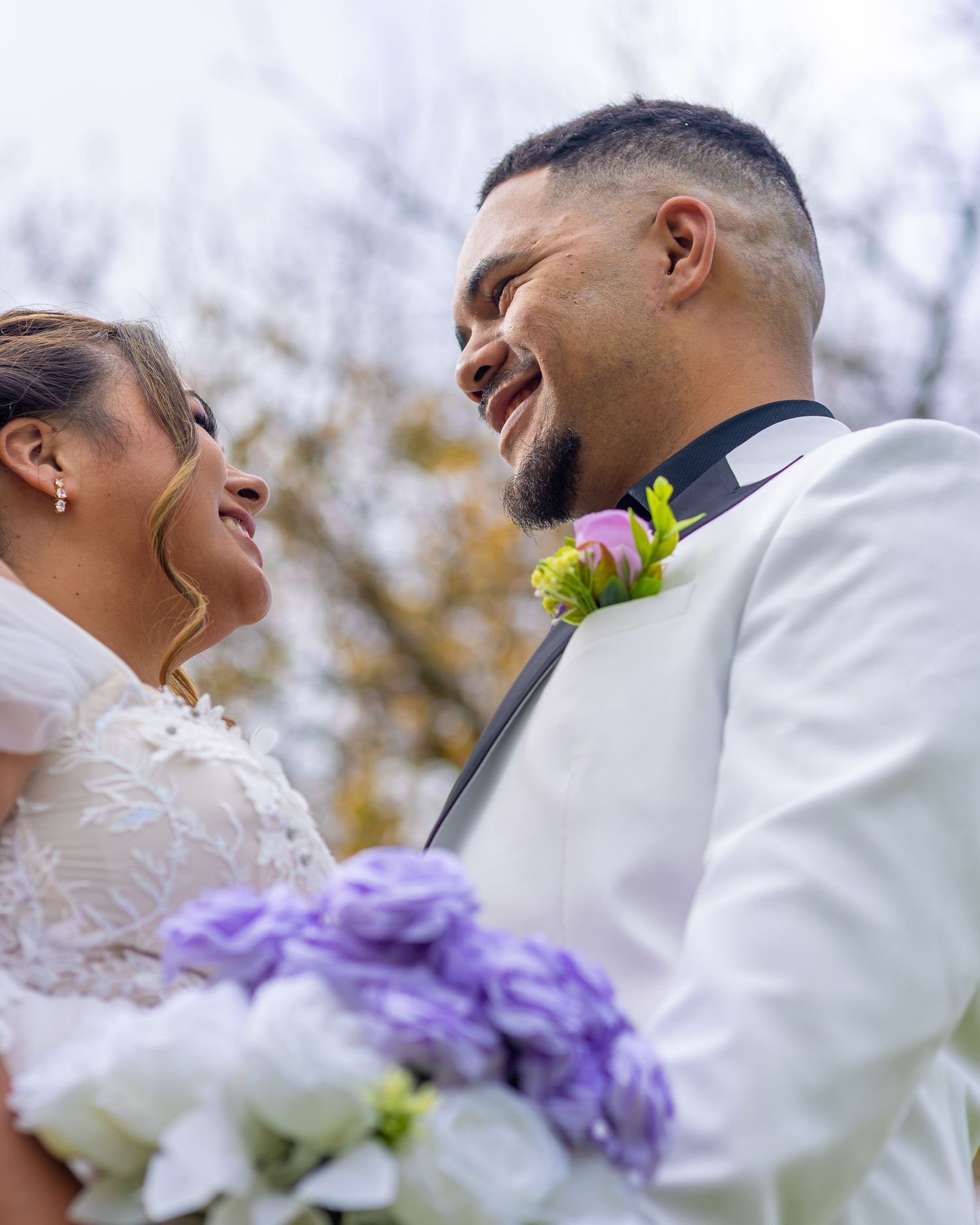 A joyful bride and groom share a loving glance outdoors, she holds purple bouquet, he wears white suit with pink boutonniere.