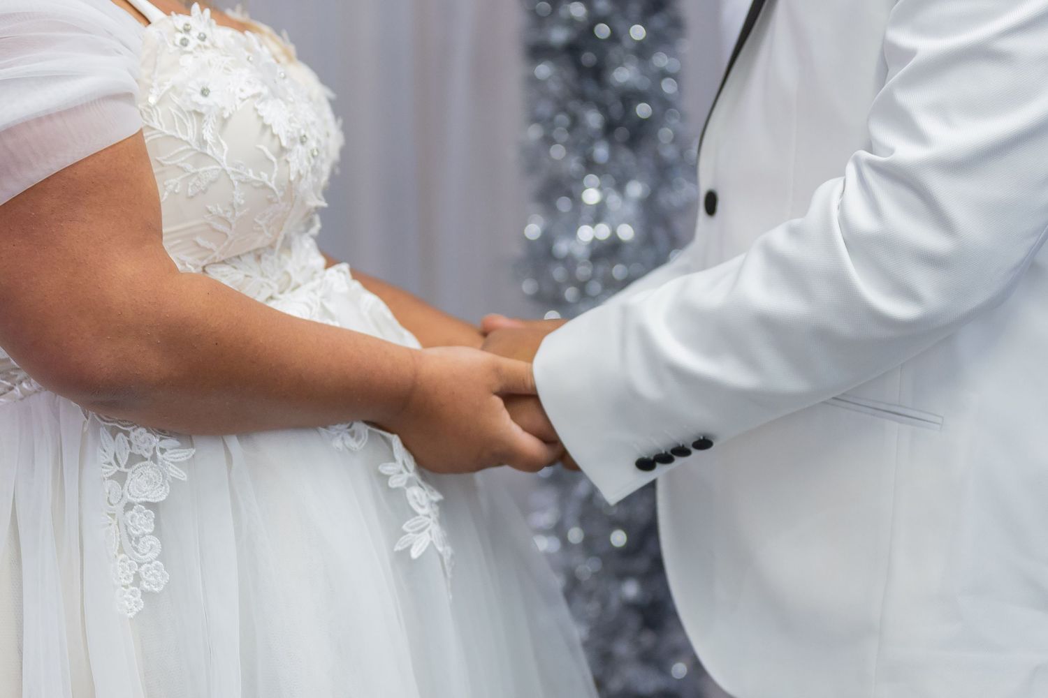 Bride and groom in white attire holding hands at wedding ceremony with sparkling backdrop.