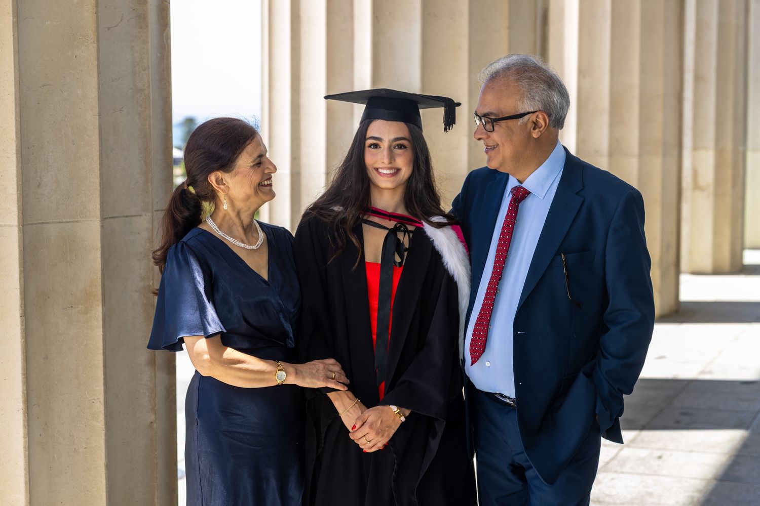 Graduate in cap and gown smiles with proud parents between stone columns at graduation ceremony.