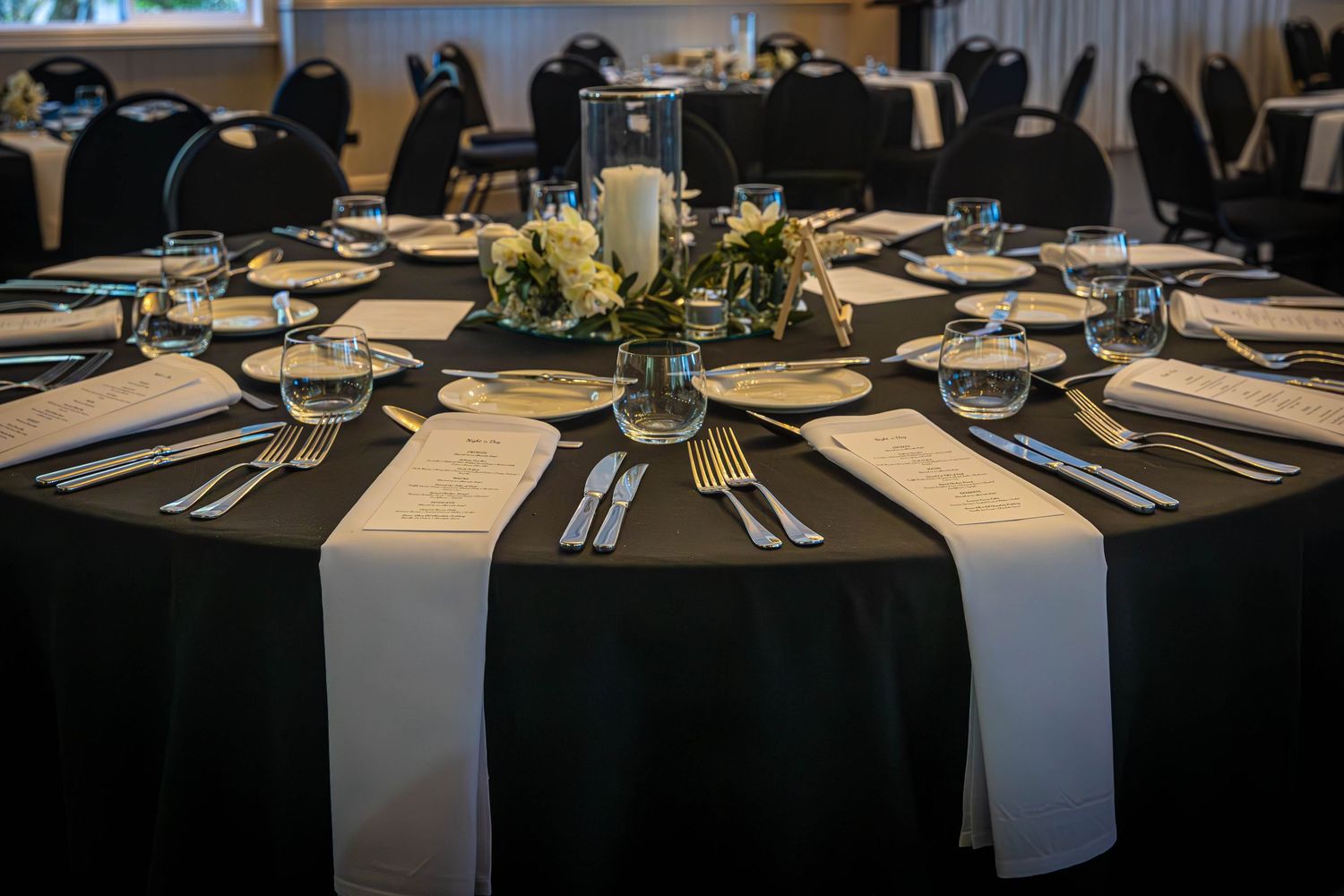 Elegant black and white banquet table setting with gold cutlery, glassware, and floral centerpiece in a formal event hall.