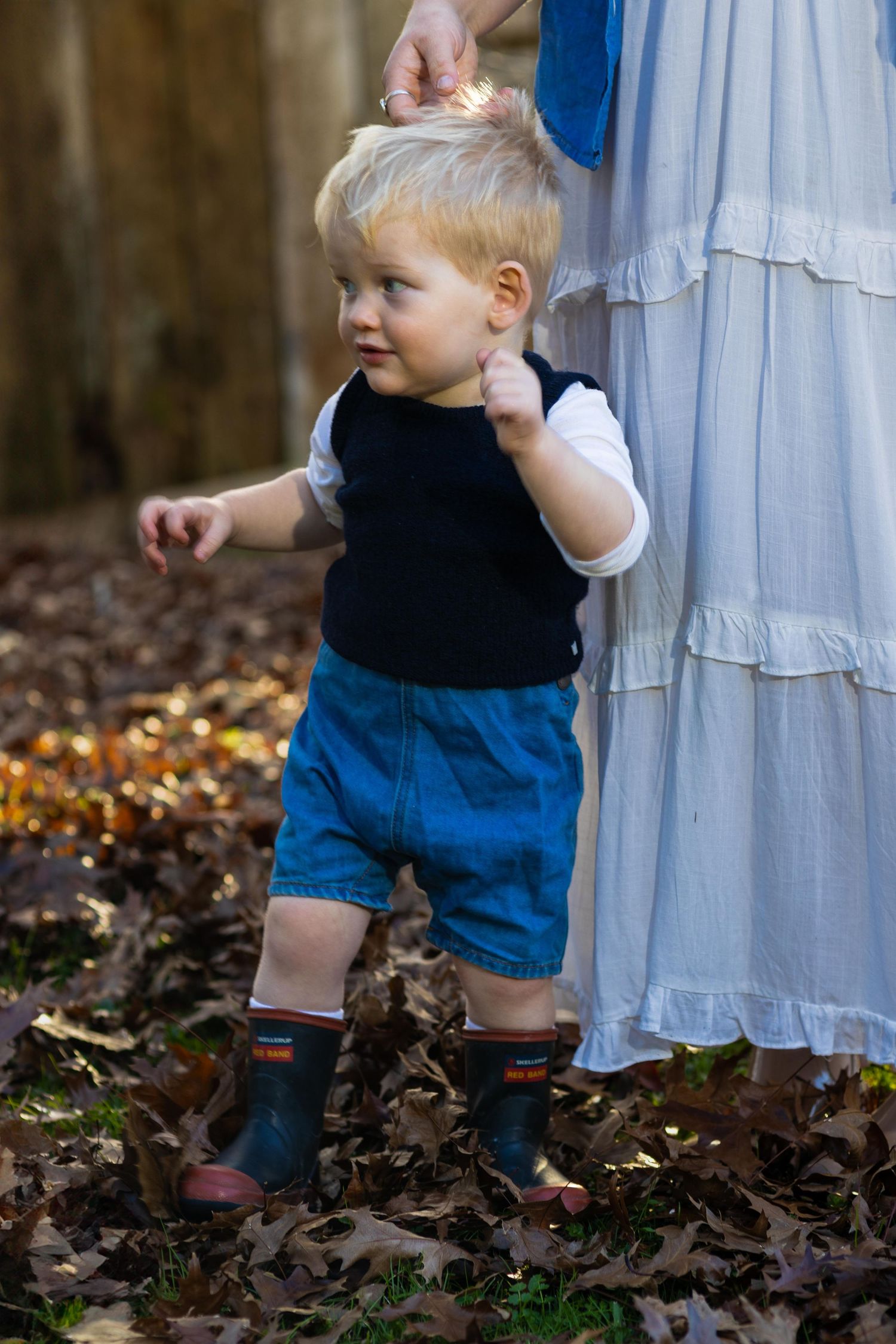 Toddler boy in denim shorts and rain boots stands in autumn leaves, holding an adult's hand outdoors.