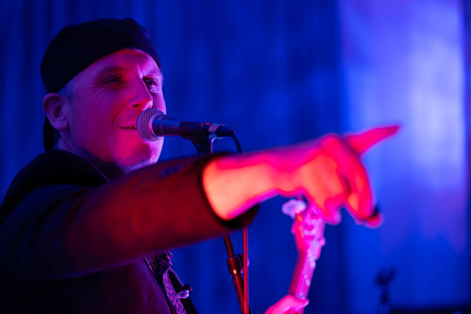 Male singer performing on stage under red and blue lights, pointing at audience near microphone.