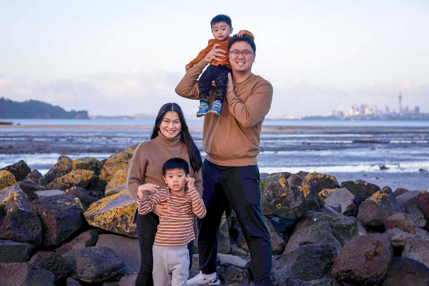 Family of four posing on rocky beach at sunset with city skyline in background.