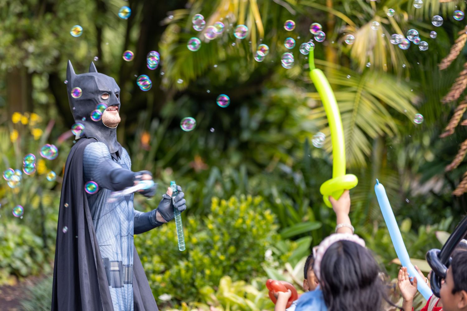 Batman character blowing bubbles with a child holding balloon animals outdoors at a theme park.