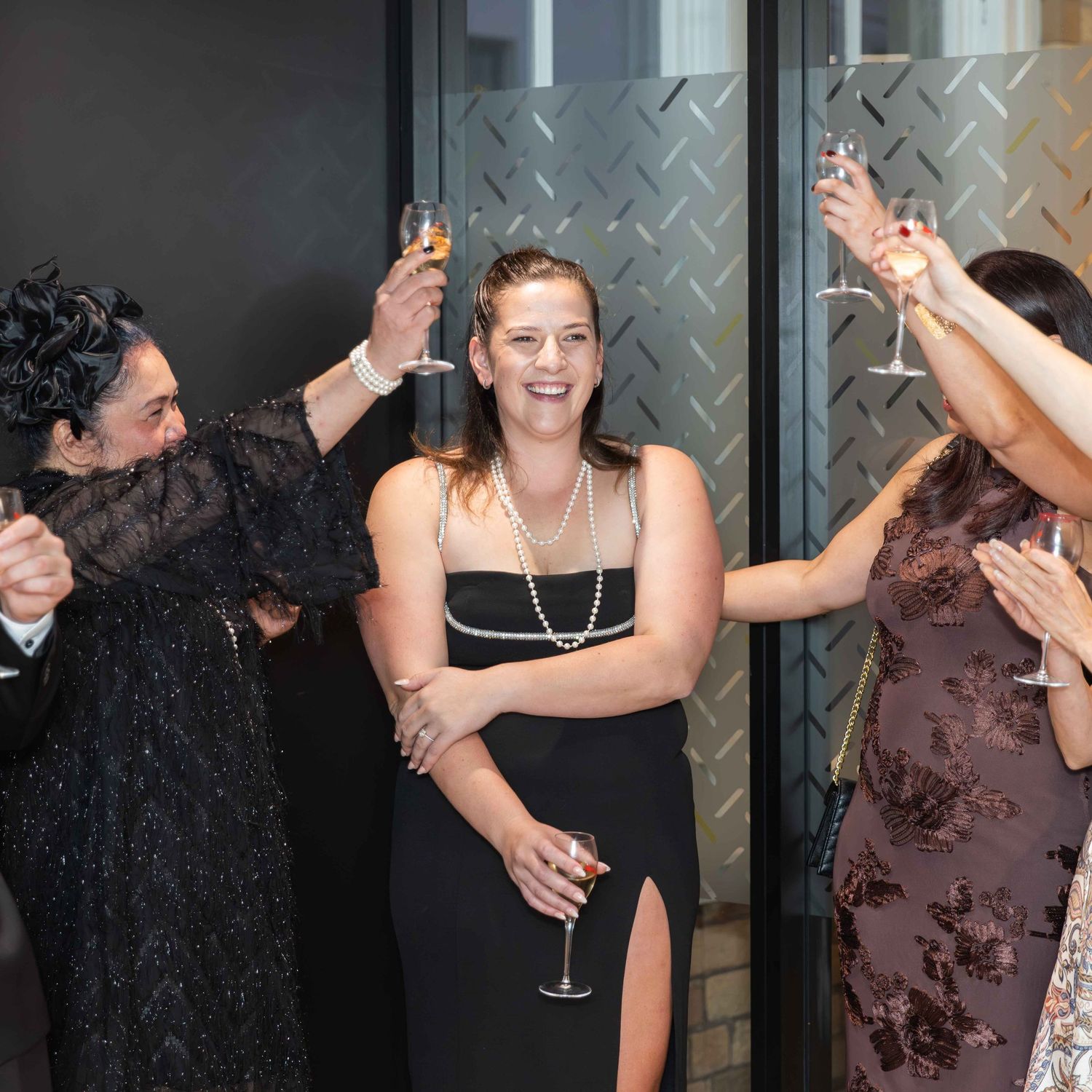 Women in elegant black gowns celebrate with champagne glasses raised at a formal evening event.
