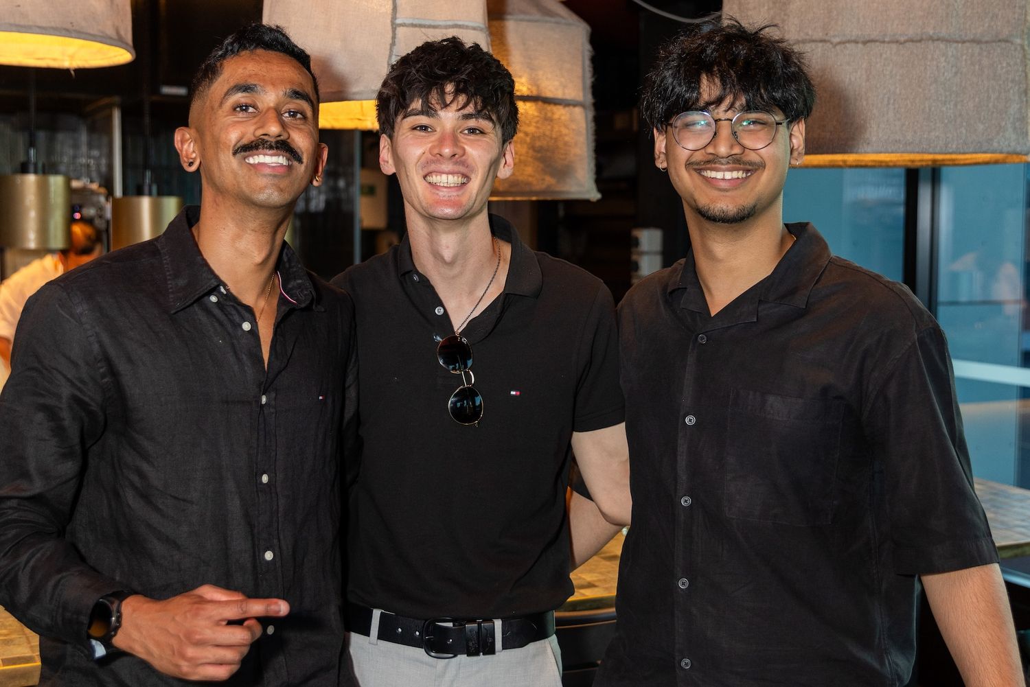 Three young men smiling together indoors, all wearing black shirts in a modern restaurant setting.