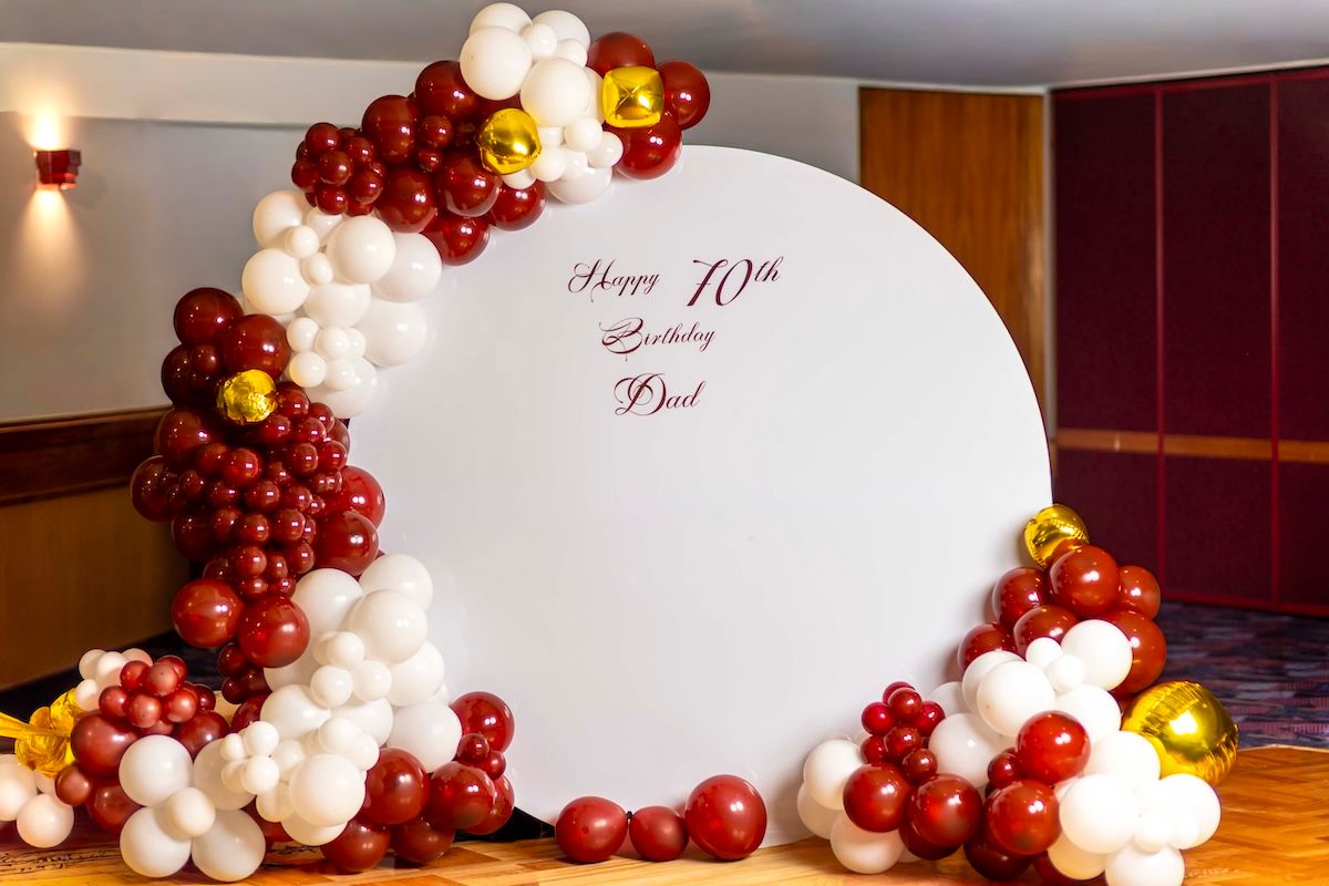 Elegant balloon arch decoration in burgundy, white and gold colors adorns an indoor event space.