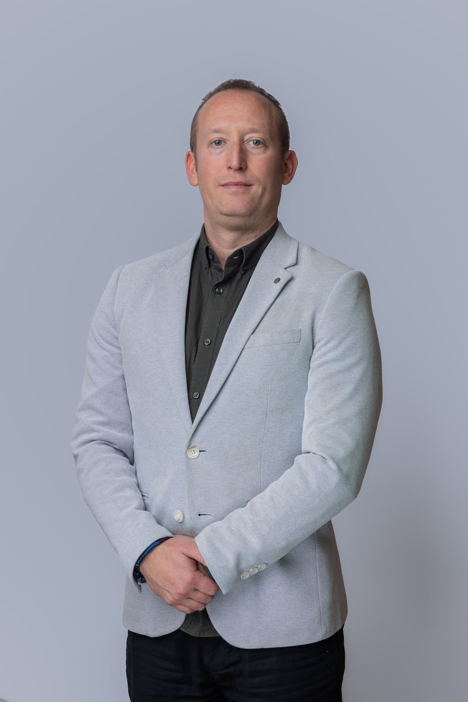 Professional man in light gray blazer and dark shirt poses confidently against a neutral gray background.