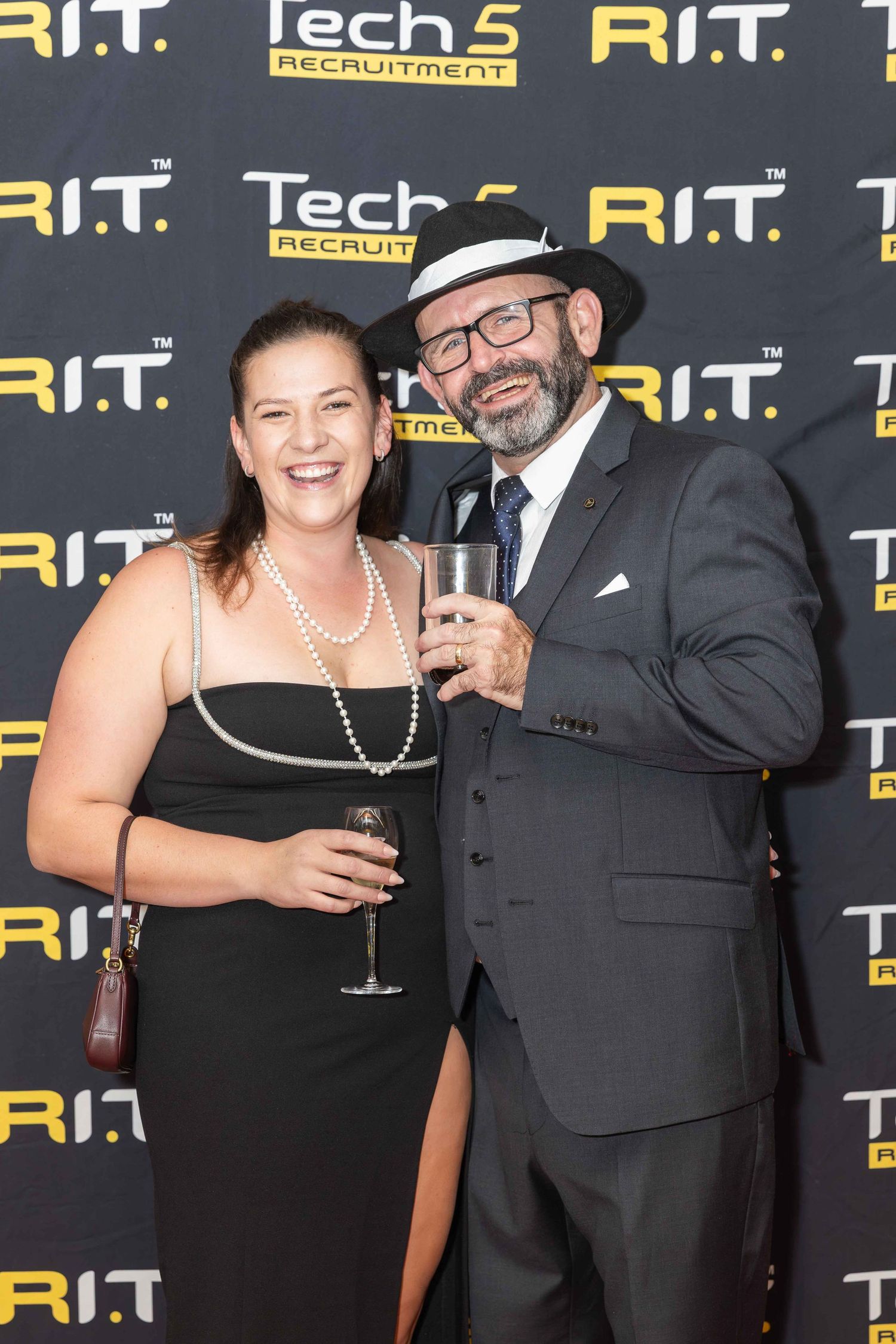 A man in a suit and hat and a woman in a black dress pose with drinks at a Tech 5 Recruitment R.I.T. event.